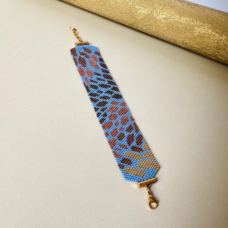 May include: A beaded bracelet with a blue background and a pattern of brown, copper, and gold beads. The bracelet has gold-colored clasps and connectors. The design is geometric and the bracelet is flat.