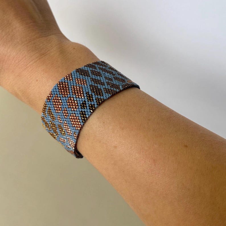 May include: A wide beaded bracelet in shades of blue, copper, and black is worn on a wrist. The bracelet features a geometric pattern of small beads. The design incorporates diamond and dot shapes, creating a textured appearance.