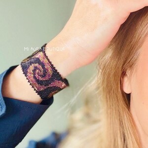 May include: A black beaded bracelet with a swirling pattern in shades of pink and gold. The bracelet is on a person's wrist.