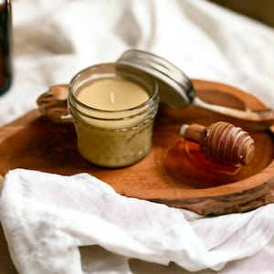 May include: A small, clear glass jar candle with a white wick sits on a wooden tray. A silver lid rests beside the candle. A wooden honey dipper and a small bottle are also on the tray. The tray is on a white cloth.