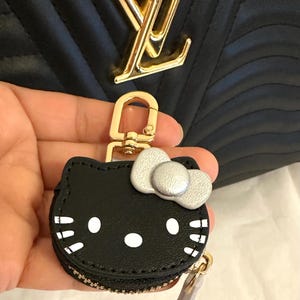May include: Black Hello Kitty coin purse with a silver bow and white accents. The purse has a gold zipper and a gold-colored clasp. The background features a black quilted handbag with a gold logo.