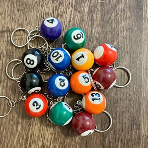 May include: A collection of billiard ball keychains featuring various colors and numbers including 1, 2, 3, 4, 5, 6, 7, 8, 10, and 13. Each small, spherical ball is attached to a silver-toned metal split ring by a short chain, displaying glossy finishes.
