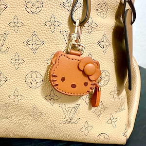 May include: A tan leather handbag with a gold-colored clasp and a brown Hello Kitty charm. The handbag has a pattern of repeating floral and logo designs. The charm is brown with a flower and a small orange tag.