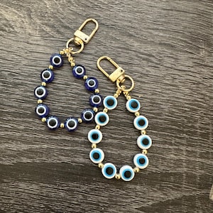 May include: Two beaded keychains featuring gold-tone metal lobster clasps. One keychain consists of dark blue evil eye beads interspersed with small gold-tone spacer beads. The second keychain features light blue and white evil eye beads.