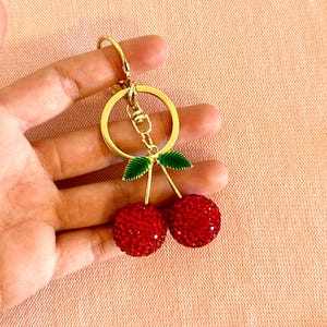 May include: A gold-colored keychain with a pair of red, crystal-studded cherries. The cherries have green leaves and are attached to a gold stem. The keychain has a gold ring and clasp.