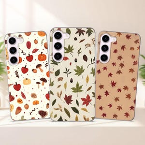 May include: Three phone cases with autumn-themed designs. One case features apples, pumpkins, and mushrooms on a cream background. Another has leaves and acorns. The third case displays scattered maple leaves.