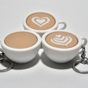 Può includere: Tre portachiavi a forma di tazza da caffè in ceramica bianca con disegni di latte art. Uno ha un cuore, un altro un motivo a foglie e il terzo una superficie di caffè liscia. Ogni portachiavi ha un anello di metallo argentato.
