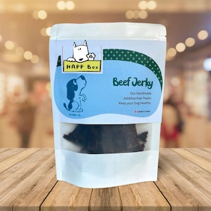 Op de afbeelding: Een witte hersluitbare zak met een blauw en groen label. Het label toont een cartoonhond en de tekst "HAPP Box Beef Jerky Our Handmade Additive-free Treats Keep your Dog Healthy Made in Canada".