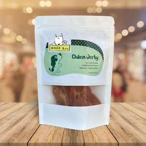 Op de afbeelding: Een witte hersluitbare zak met een groen-wit etiket. Het etiket toont een cartoonhond en de tekst "Happ Box Chicken Jerky Our Handmade Additive-free Treats Keep your Dog Healthy". De zak bevat een paar stukjes gedroogde kip.