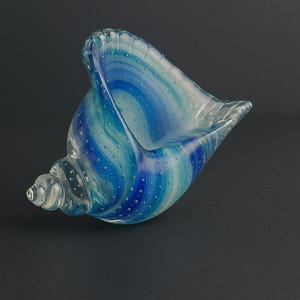 May include: A decorative glass sculpture shaped like a conch shell, featuring swirling bands of translucent blue, teal, and white glass. The surface is embedded with numerous small, clear air bubbles throughout the material, creating a textured effect.