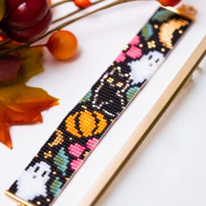 May include: A rectangular beaded bracelet featuring a black background with a repeating pattern of white ghosts, orange pumpkins, pink flowers, and green leaves. The bracelet is finished with gold-toned metal end bars at both ends of the beaded section.