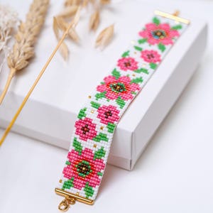 May include: A beaded bracelet with a floral design of pink flowers and green leaves on a white base. The bracelet has gold-coloured clasps and accents. The flowers have a dark brown centre. The bracelet is on a white surface.