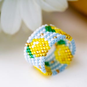 May include: A wide band ring crafted from small glass seed beads in a woven pattern. The design features yellow and green pineapple motifs set against a light blue and white background, accented with metallic gold beads throughout the structure.