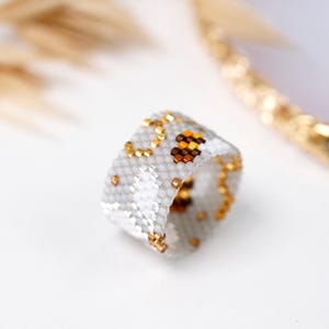Può includere: Un anello a fascia larga realizzato con piccole perline opache color grigio chiaro. L'anello presenta un motivo ripetitivo di minuscole perline metalliche dorate e marroni disposte in forme astratte e piccoli gruppi sulla superficie grigia.