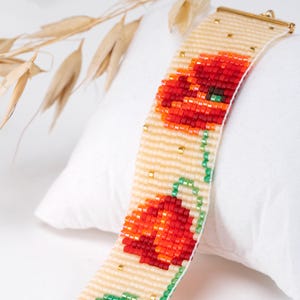 Könnte beinhalten: Ein Armband mit Perlen mit einem Mohnblumenmuster. Das Armband hat einen cremefarbenen Hintergrund mit roten und orangen Mohnblumen und grünen Stielen. Goldfarbene Perlen sind eingestreut, und der Verschluss ist goldfarben.