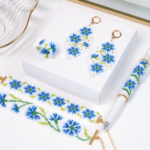 Peut inclure: Un ensemble de bijoux blancs avec broderie florale bleue et verte. L'ensemble comprend une bague, des boucles d'oreilles, un bracelet et un stylo. Les boucles d'oreilles sont en forme de losange avec des cerceaux dorés. Le bracelet a un fermoir doré.