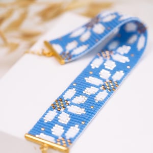 May include: A wide beaded bracelet featuring a repeating floral pattern in light blue, white, and metallic gold seed beads. The bracelet is finished with gold-toned metal end bars and a matching clasp, creating a structured, flat band design.