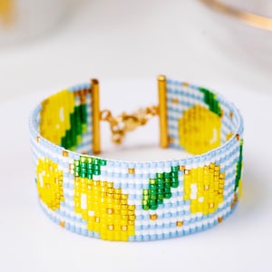 May include: A beaded bracelet featuring a light blue and white gingham pattern with yellow lemon motifs and green leaves. The bracelet is constructed from small square beads and includes gold-toned metal end bars and a gold-toned lobster claw clasp.