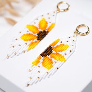 May include: A pair of beaded earrings with a sunflower design. The earrings have a white background with yellow and brown sunflower details. The fringe is white with gold accents, and the hoops are gold.