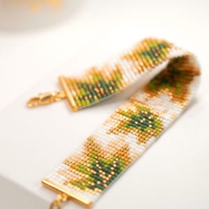 May include: A wide beaded bracelet featuring a repeating geometric pattern in shades of white, olive green, and metallic gold. The bracelet is constructed from small square beads and finished with gold-toned metal end bars and a lobster claw clasp.