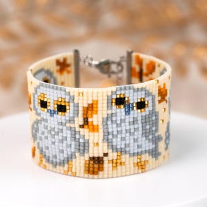 May include: A wide cuff bracelet crafted from small beads in a cream color, featuring a repeating pattern of two grey owls with black and gold eyes. The design includes scattered orange and brown leaf-like shapes and a silver-toned metal clasp.