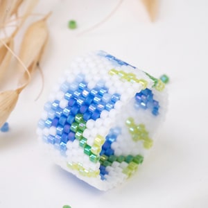 May include: A handmade beaded ring featuring a floral pattern in white, blue, and green. The ring is cylindrical and appears to be made of small, colorful beads. The design incorporates shades of blue and green against a white background, creating a textured, decorative accessory.