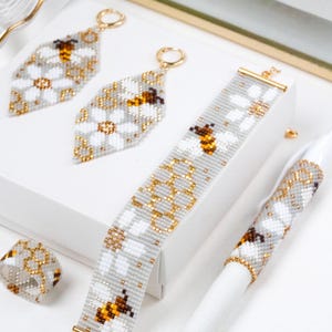 Pode incluir: Um conjunto de joias e acessórios de missangas com um padrão repetitivo de abelhas, margaridas brancas e motivos de favos de mel dourados sobre um fundo cinzento claro. O conjunto inclui dois brincos em forma de diamante, uma pulseira, um anel e uma capa de caneta.