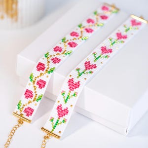 Peut inclure: Deux bracelets blancs perlés avec des motifs floraux roses et des accents dorés. Les bracelets présentent un motif répété de fleurs roses avec des feuilles vertes et des perles dorées. Chaque bracelet a un fermoir et une chaîne dorés.