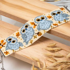 May include: A wide beaded cuff bracelet featuring a repeating pattern of light blue owls with black eyes and orange accents on a cream background. The bracelet is finished with silver-toned metal end bars and a delicate silver-toned chain extender.