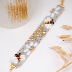 May include: A beaded bracelet featuring a light gray background with a repeating pattern of white and brown bees, gold honeycomb shapes, and white flowers. The bracelet is finished with gold-toned metal end bars and a small gold-toned clasp.