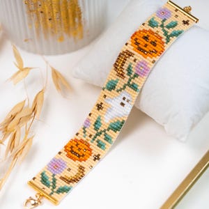 May include: A wide beaded bracelet featuring a cream-coloured background with a repeating pattern of orange jack-o'-lanterns, white ghosts, purple flowers, green leaves, and brown crescent moons, finished with gold-toned metal end bars and a clasp.