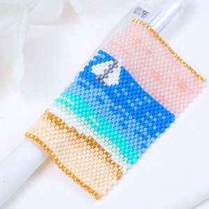 May include: A beaded pen wrap in shades of blue, white, and peach, with a gold border. The wrap features a gradient design and a small, pixelated heart. The pen wrap is made of small, colourful beads.