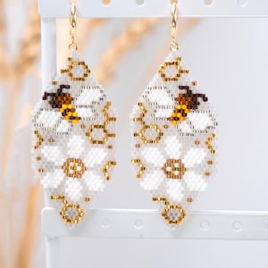 Könnte beinhalten: Ein Paar rautenförmige Perlenohrringe mit einem weißen Gänseblümchenmuster und einer braun-gelben Biene in der Mitte. Das Design besteht aus kleinen weißen, goldenen, braunen und gelben Perlen, die an goldfarbenen Metall-Creolen befestigt sind.