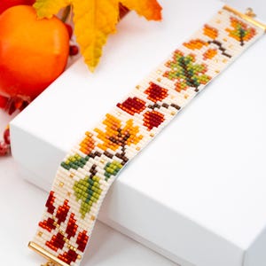 May include: A wide beaded bracelet featuring a cream-colored background with a repeating pattern of autumn leaves in shades of red, orange, yellow, and green. The bracelet is finished with gold-toned metal end caps at both the top and bottom edges.