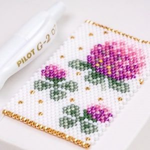 May include: A rectangular beaded patch featuring a white background with a floral pattern of pink and purple blossoms and green leaves. The design includes small gold accent beads and a gold beaded border. A white Pilot G-2 07 pen is visible.
