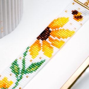 May include: A beaded bracelet featuring a sunflower and bee design. The bracelet is primarily white with yellow, orange, brown, and green beads. The sunflower has yellow petals and a brown center. A small bee is near the top. The bracelet is on a white surface.