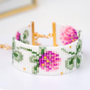 May include: White beaded bracelet featuring a floral design with pink, purple, green, and yellow accents. The bracelet has a gold-colored clasp and is approximately 1.5 inches wide.