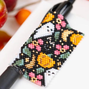 Op de afbeelding: Een zwarte pen met een kralenhoes met een patroon van witte geesten, oranje pompoenen, roze bloemen en groene bladeren. Het ingewikkelde kralenwerk bedekt de houder van de pen en toont een gedetailleerd seizoensontwerp met kleine metalen accenten.