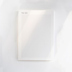 Könnte beinhalten: Ein rechteckiger weißer Notizblock mit einem Raster aus dünnen grauen Linien und dem Text TO DO, der in Schwarz in der oberen linken Ecke gedruckt ist. Das Papier hat eine saubere, glatte Oberfläche mit einem einheitlichen Layout.