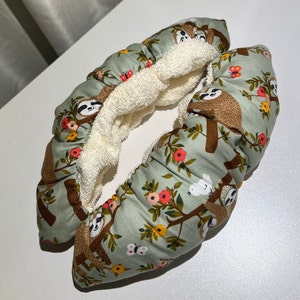 May include: A pair of green and white skate guards with a sloth and floral pattern. The guards are lined with white fleece.