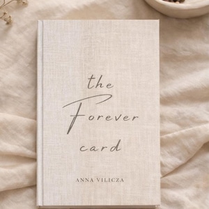 May include: A hardback book with a linen-textured cover. The title "the Forever card" is written in elegant script, with the author's name, Anna Vilicza, below. The book is a neutral beige colour.