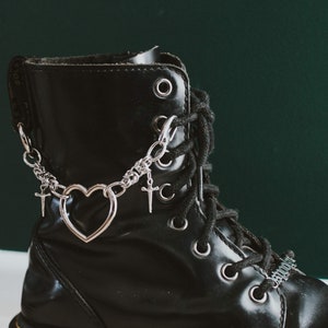 Puede incluir: Primer plano de una bota de cuero negro con detalles de cadena plateada. La cadena presenta dijes de corazón y cruz. La bota tiene una suela de plataforma y cordones negros. El fondo es verde oscuro.