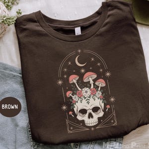 May include: Brown t-shirt featuring a skull with mushrooms, flowers, and a crescent moon design. The graphic includes stars and a decorative arch. The shirt is folded and displayed on a white surface.