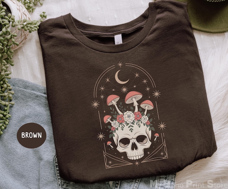 May include: Brown t-shirt featuring a skull with mushrooms, flowers, and a crescent moon design. The graphic includes stars and a decorative arch. The shirt is folded and displayed on a white surface.