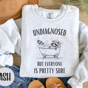 Może przedstawiać: Szara bluza z nadrukiem  rakuma w wannie i tekstem "Undiagnosed but everyone is pretty sure". Bluza leży na niebieskim dżinsie.