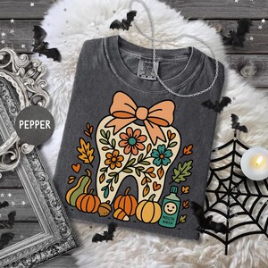 May include: A dark gray t-shirt featuring a whimsical tooth design with a bow, flowers, and pumpkins. The shirt has a vintage wash look and includes the text "Mouth Wash" on a small bottle. Halloween-themed apparel.