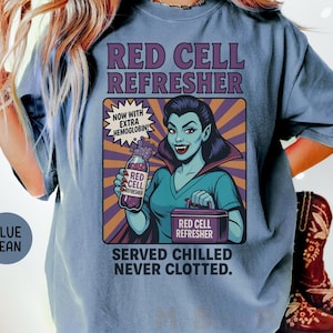 May include: Blue jean t-shirt featuring a retro-style graphic. The graphic depicts a vampire-like character holding a bottle labeled "Red Cell Refresher." Text includes "Now with extra hemoglobin!" and "Served chilled, never clotted."