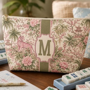 May include: A zippered pouch featuring a cream-colored fabric with a pink tiger, floral patterns, and pagoda illustrations. A central olive green vertical stripe displays a decorative frame containing the capital letter M in olive green.