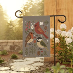 May include: A decorative garden flag featuring three illustrated birds: a red cardinal, a brown robin, and a green and white chickadee. The flag has a gray background with floral and snowflake accents. The flag is displayed on a black metal stand.