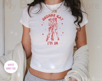 May include: A white short-sleeved baby tee featuring a red graphic print of two figures in cowboy hats and boots standing together. The text "SOUNDS GAY I'M IN" is printed in red above and below the illustration, accented by small red stars.